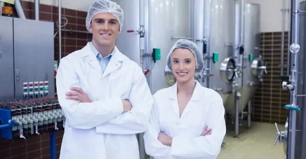 Two technicians in lab coats and hairnets standing before stainless process vessels